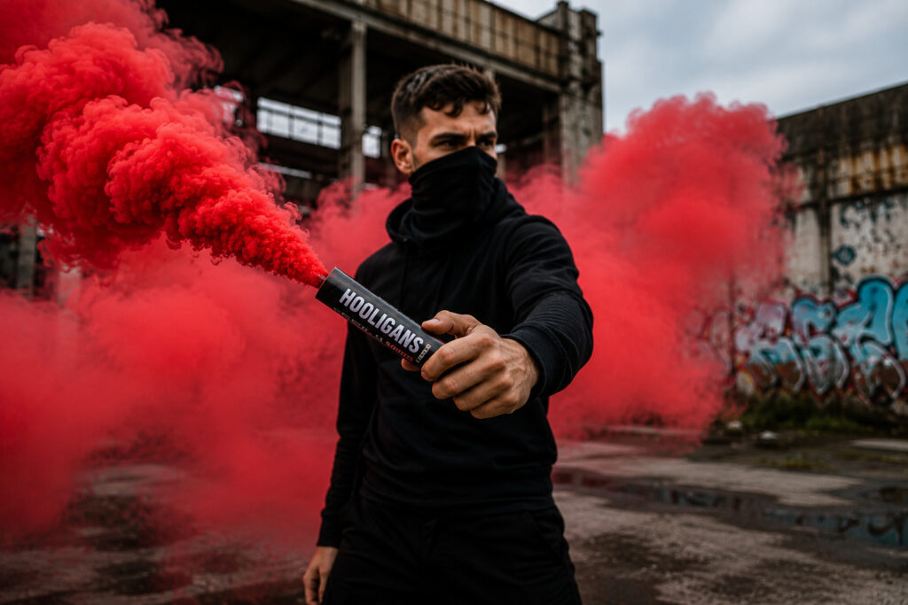 Mężczyzna trzymający świecę dymną do ręki Hooligans HDP60 emitującą intensywny czerwony dym w industrialnym otoczeniu