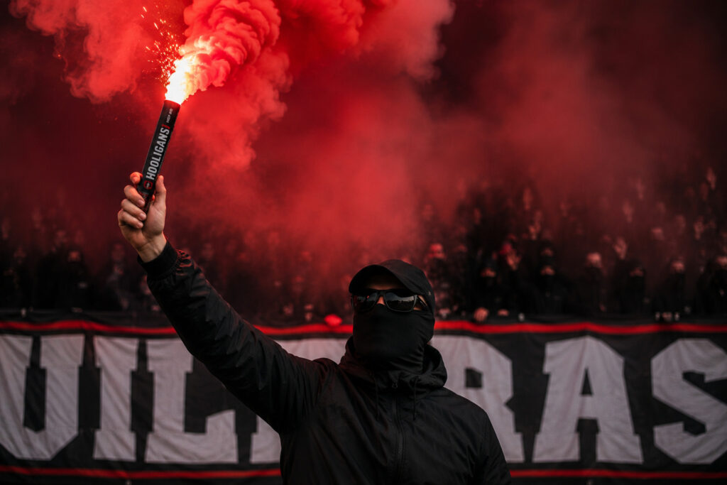 Kibic trzymający świecę dymną do ręki podczas oprawy stadionowej z czerwonym dymem
