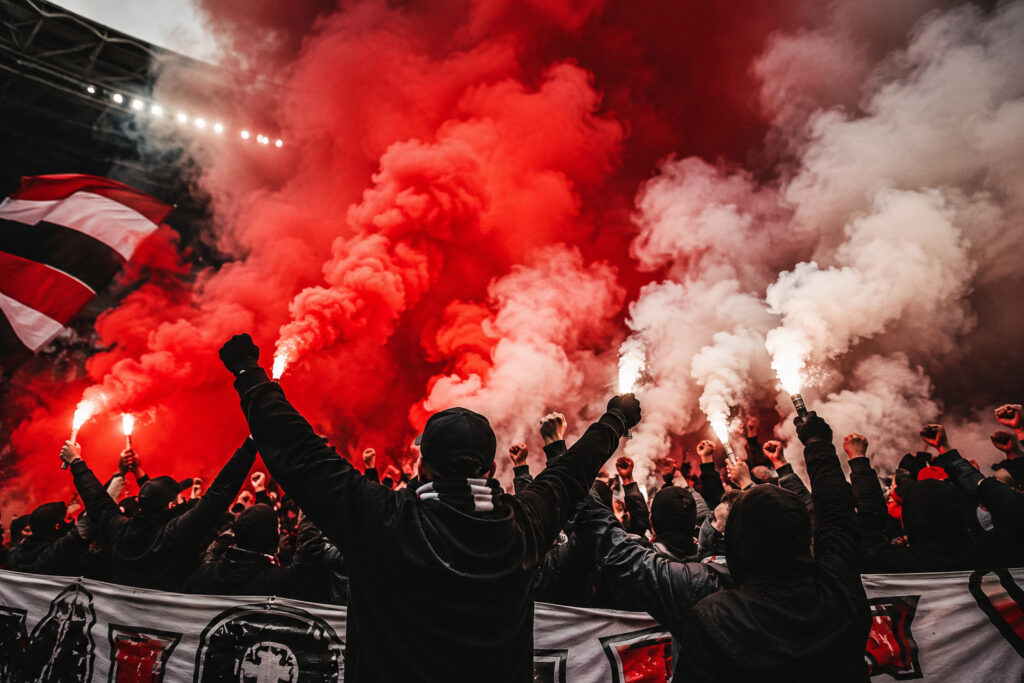 Kibice na stadionie z czerwonymi i białymi świecami dymnymi podczas oprawy meczowej