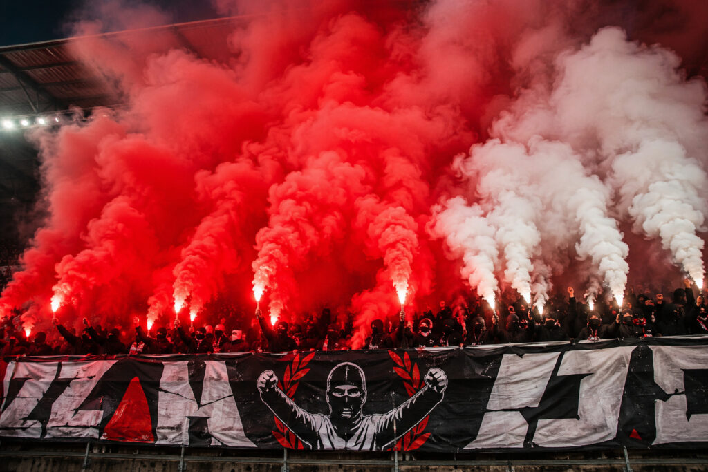 Kibice prezentujący oprawę stadionową z wykorzystaniem czerwonych i białych świec dymnych